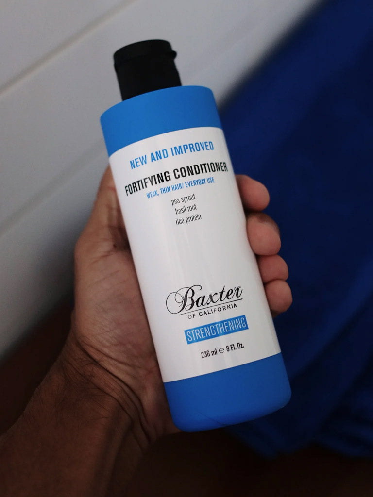 A hand holds Baxter Fortifying Conditioner, a blue and white bottle labeled for weak, thin hair and daily use. This thickening formula with pea sprout, basil root, and rice protein helps strengthen and volumize hair.
