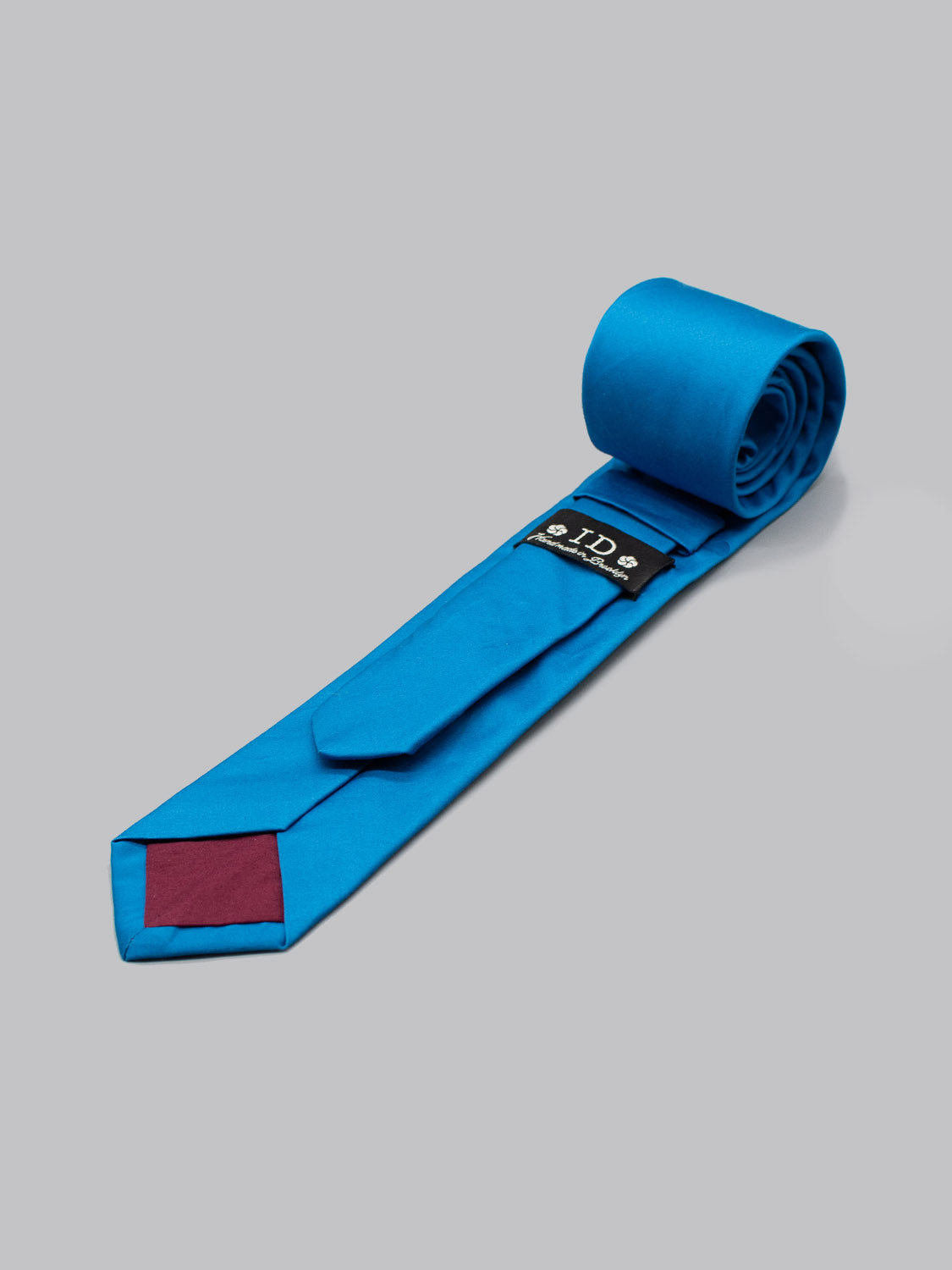 A Blue Jay Cotton Tie by ID Menswear lies unrolled on a gray backdrop, featuring a maroon tip patch and a black label with text and symbols near the narrow end. Handmade in Brooklyn from 100% cotton.