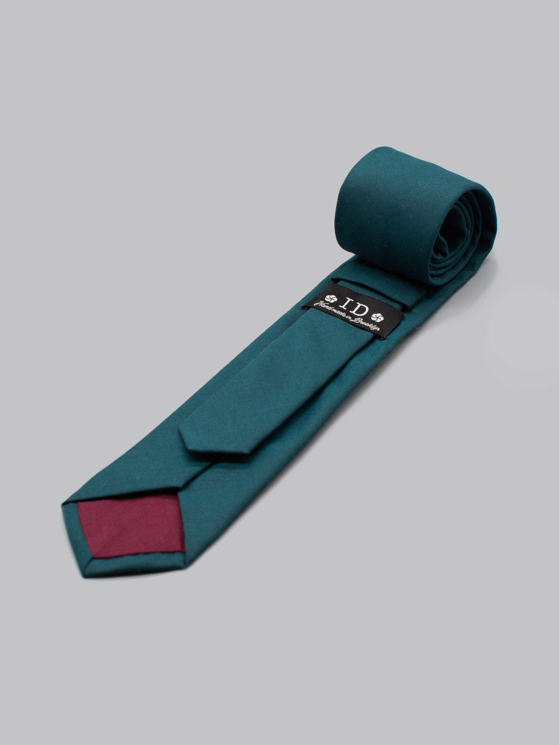 The Emerald Cotton Tie by ID Menswear, featuring a maroon tip and black label with white text and skull icons, is handmade in Brooklyn from cotton and displayed rolled on one end against a light gray background—ideal for business formal wear.