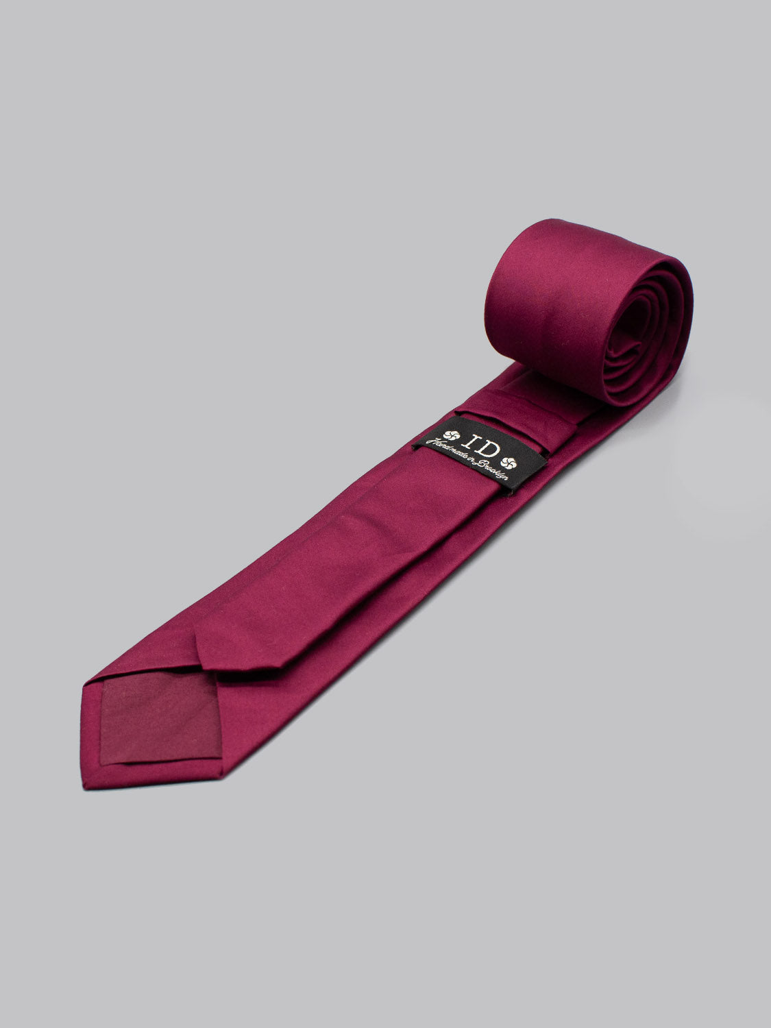A rolled, solid magenta Egyptian Firefinch Cotton Tie by ID Menswear rests on a light gray surface, featuring a black label near the base—handmade in Brooklyn and ideal for business formal occasions.