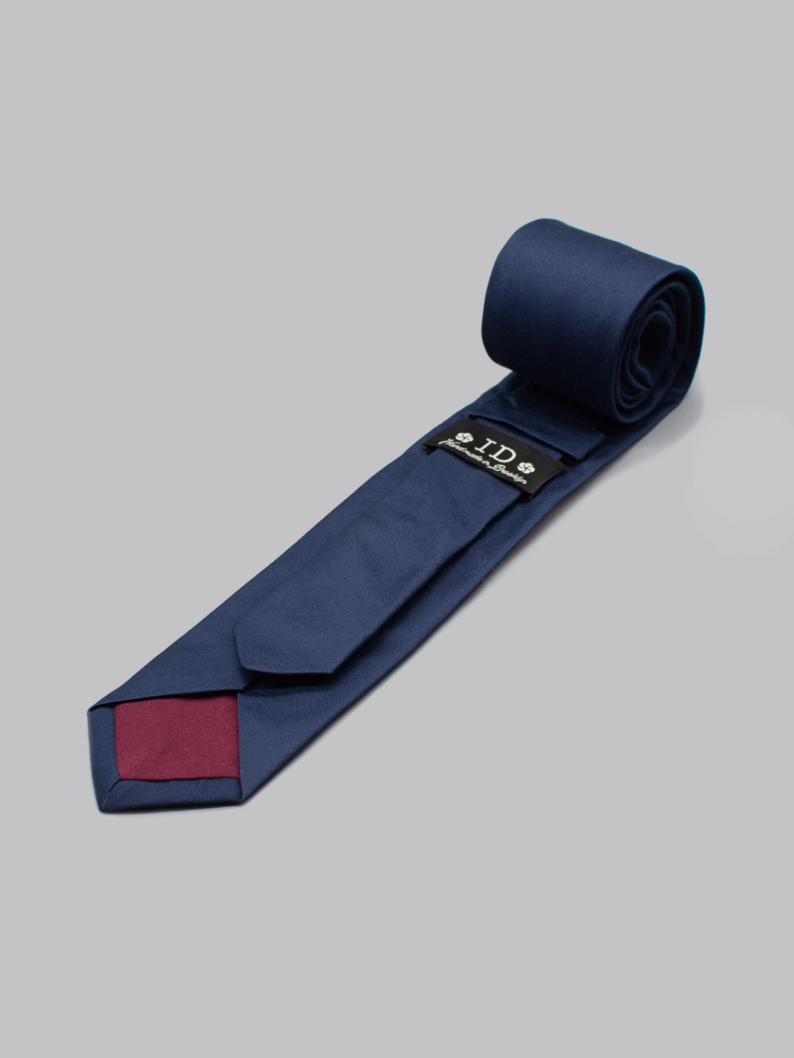 The Indigo Cotton Tie by ID Menswear, handmade in Brooklyn, features a rolled end revealing a maroon tip and black label with white text, set on a plain light gray background—ideal for business formal attire.