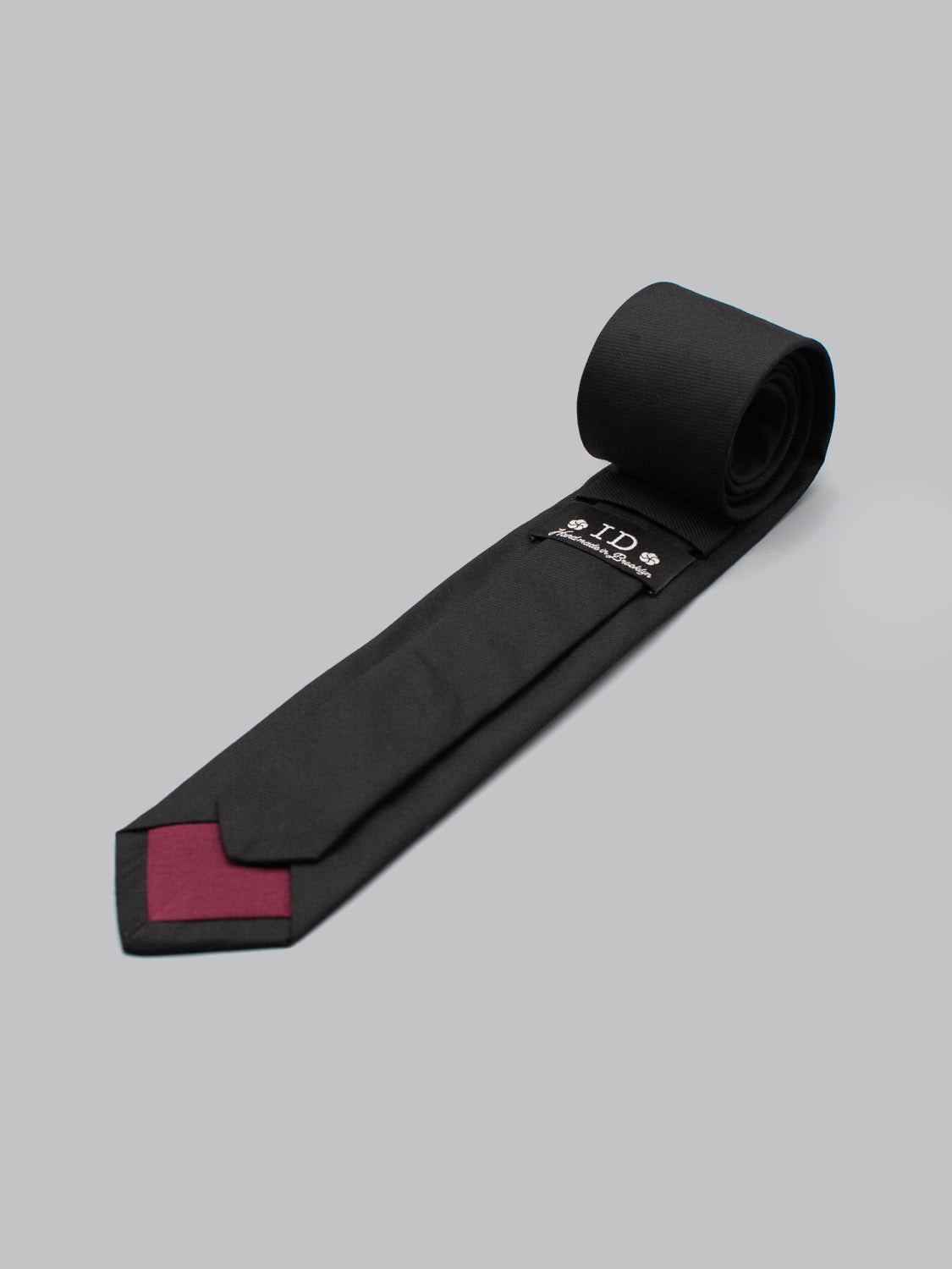A rolled-up Raven Black Cotton Tie by ID Menswear with a burgundy tip rests on a light gray background. Handmade in Brooklyn from 100% cotton, its underside shows a small white embroidered logo and text.