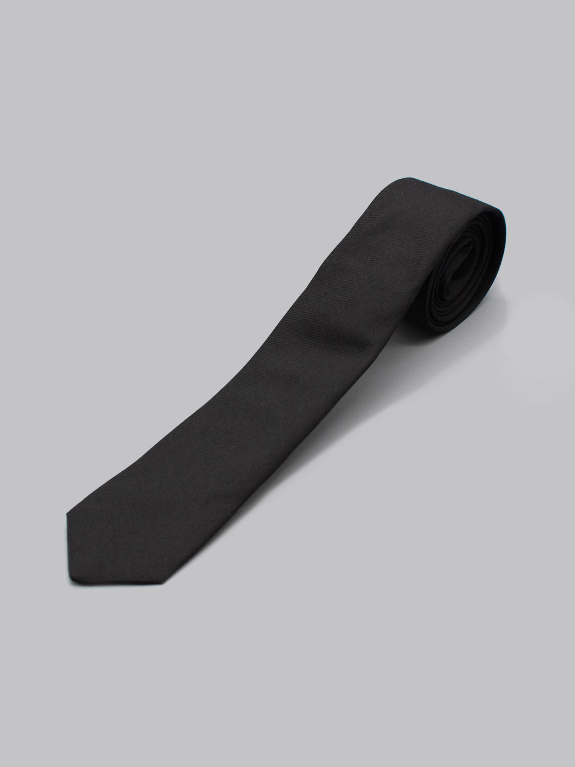 The Skinny Raven Black Cotton Tie by ID Menswear is handmade in Brooklyn from 100% cotton, shown rolled at one end on a plain light gray background.