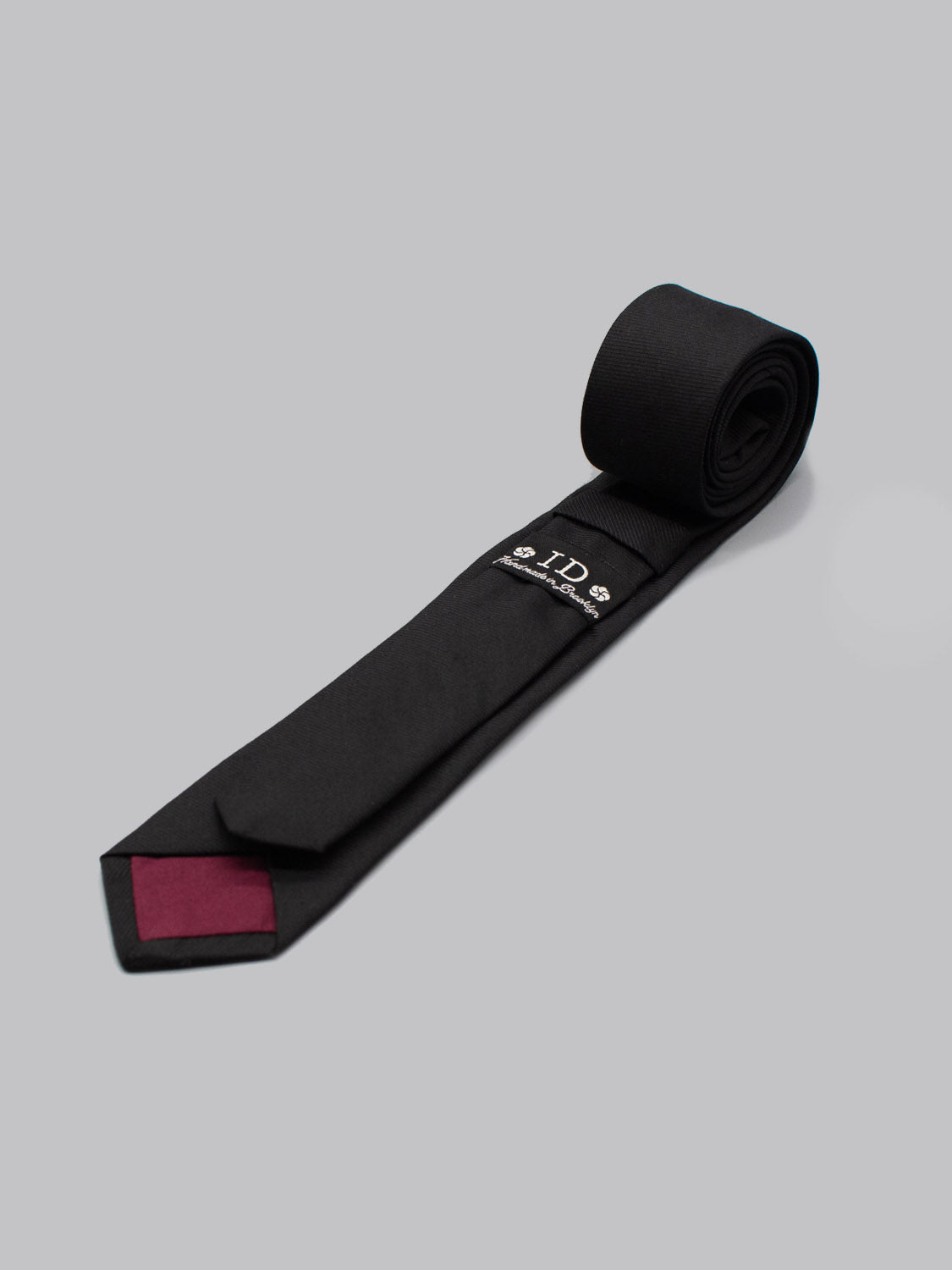 The Skinny Raven Black Cotton Tie by ID Menswear is a handmade, 100% cotton skinny tie in black, featuring a maroon inner tip and embroidered label, shown neatly rolled on a light gray background.