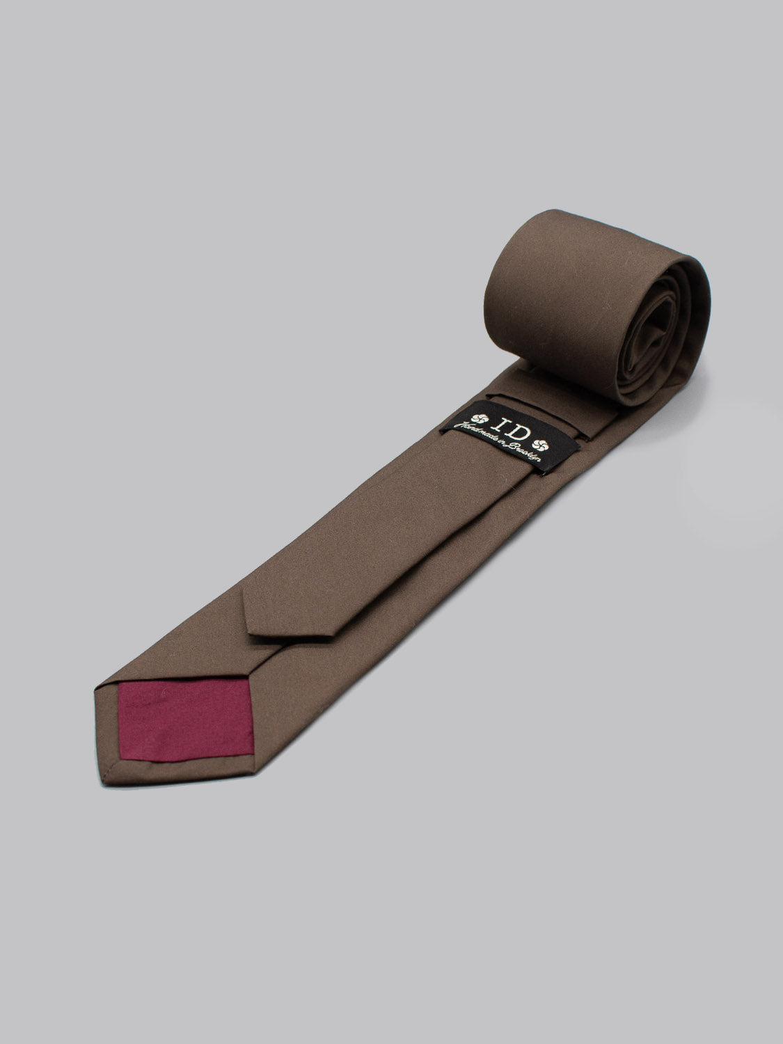 The Sparrow Cotton Tie by ID Menswear is a rolled brown cotton tie with a maroon tip, shown on a light gray background. The white-logo label highlights its handmade quality in Brooklyn, making it a refined business formal accessory.