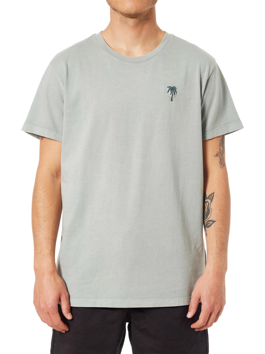 Wearing the Katin Crawford Embroidered Tee with a subtle palm tree design, someone stands out against a plain background. This light gray, organic cotton shirt from Katin complements tattooed arms and dark pants, capturing a minimalist sunset vibe.