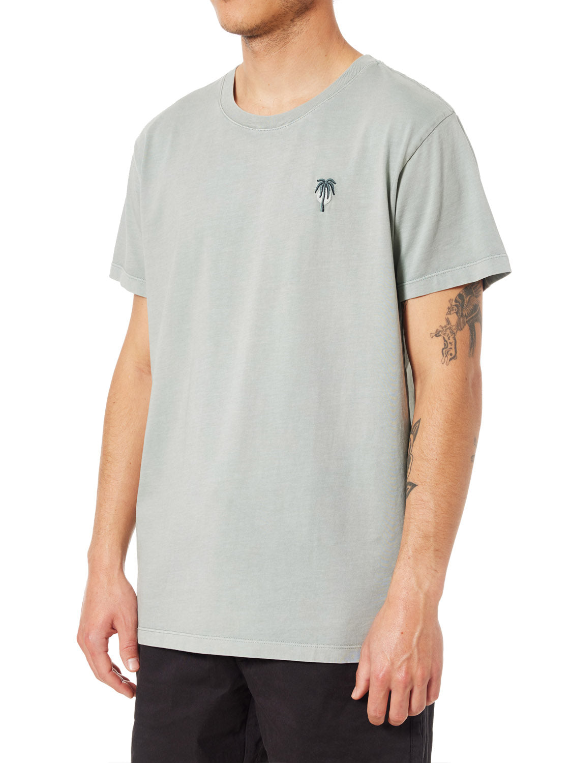 A person is seen sporting Katins light green Crawford Embroidered Tee, featuring a small palm tree logo on the chest. Made from organic cotton, it pairs well with their black pants and forearm tattoos. Their head is not visible in the frame.