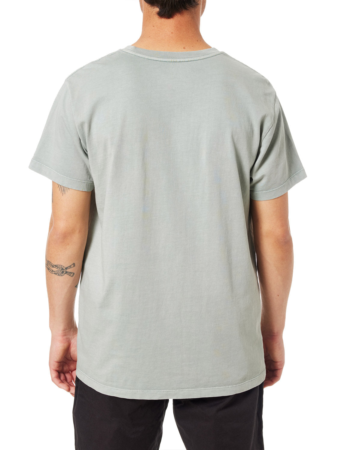 Facing away from the camera, a person wears the Katin Crawford Embroidered Tee made of organic cotton with black pants. Against the plain white backdrop, a small tattoo on their left arm echoes a minimalist style reminiscent of sunset silhouettes.