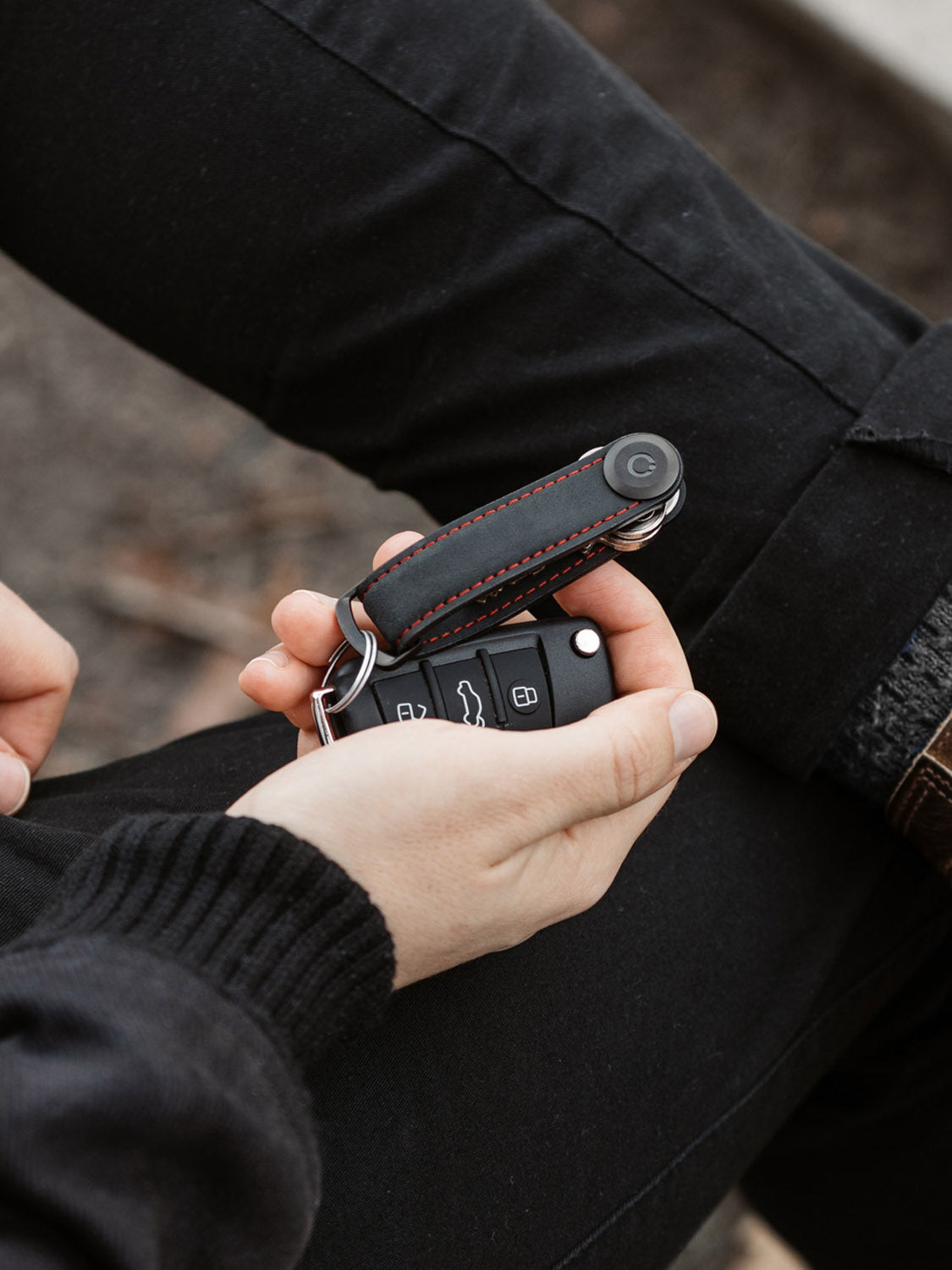 A person in black pants holds a set of car keys and a multitool with an Orbitkeys Key Organizer Crazy-Horse Cowhide Leather, featuring red stitching, in their hands.