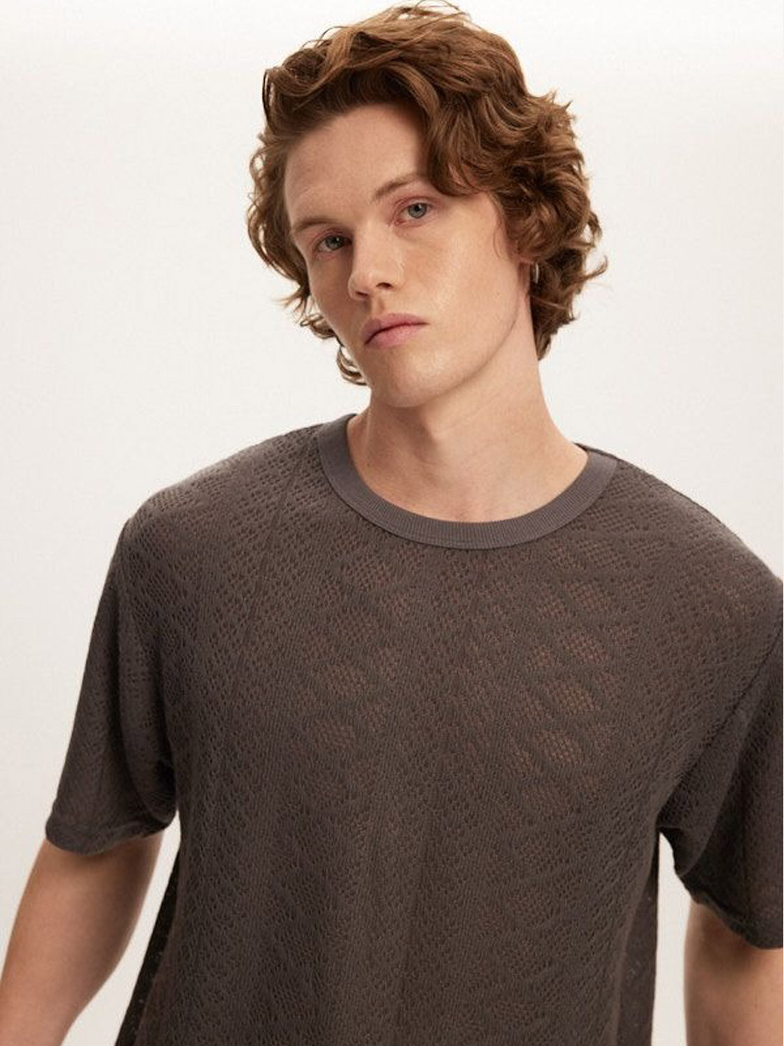 A person with wavy brown hair wears the Kuwalla Crochet Jersey Tee, a classic fit in dark gray with a crochet texture, looking slightly to the side against a simple light background.