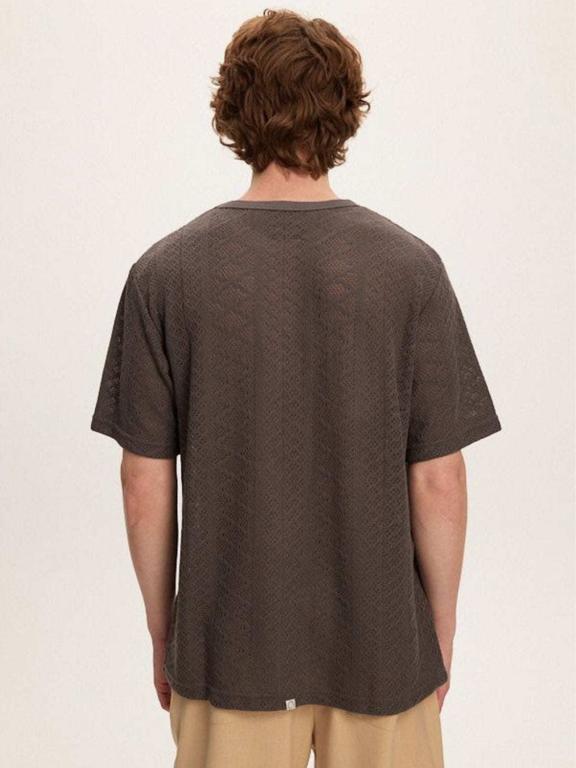 Rear view of a person with curly hair wearing the Kuwalla Crochet Jersey Tee, a gray crochet-textured short-sleeve shirt, paired with classic fit beige pants, standing against a light background.