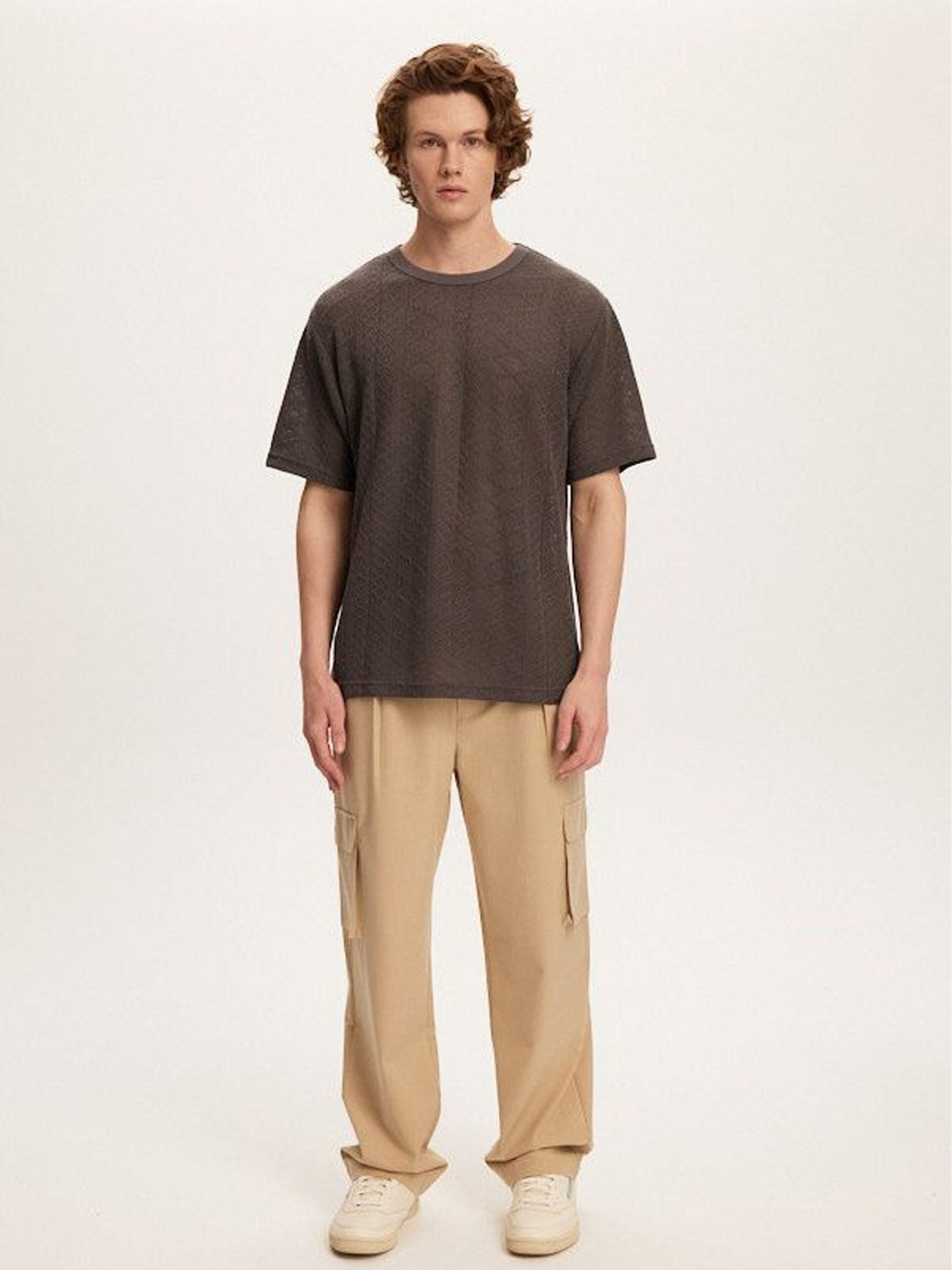 A person with curly hair stands against a plain background, wearing a Kuwalla Crochet Jersey Tee in a classic fit and light-colored cargo pants. The soft, breathable fabric complements their neutral expression as they gaze directly at the camera.