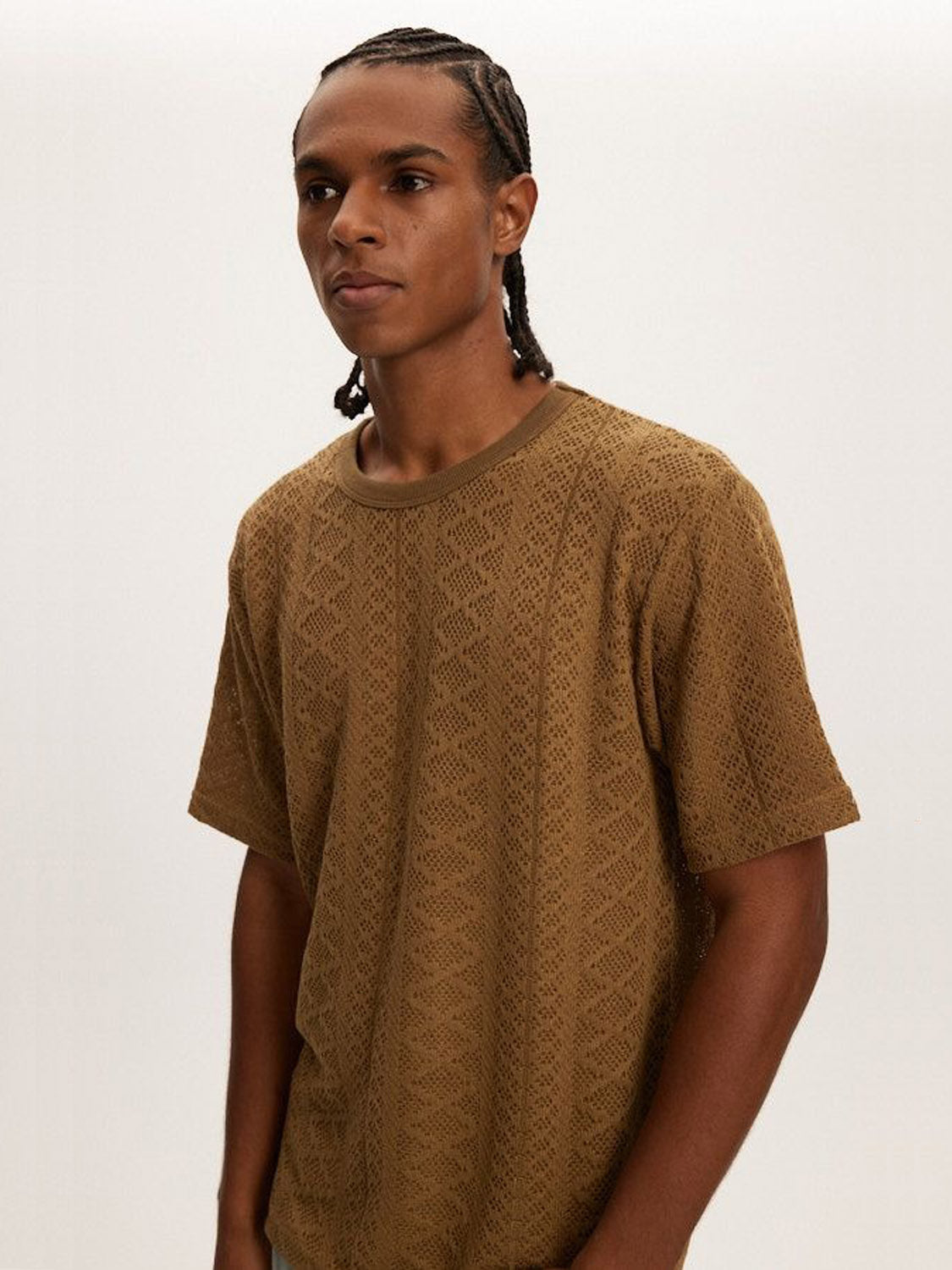 A person with braided hair, wearing a Kuwalla Crochet Jersey Tee in classic fit, stands against a plain white background, looking to the side.