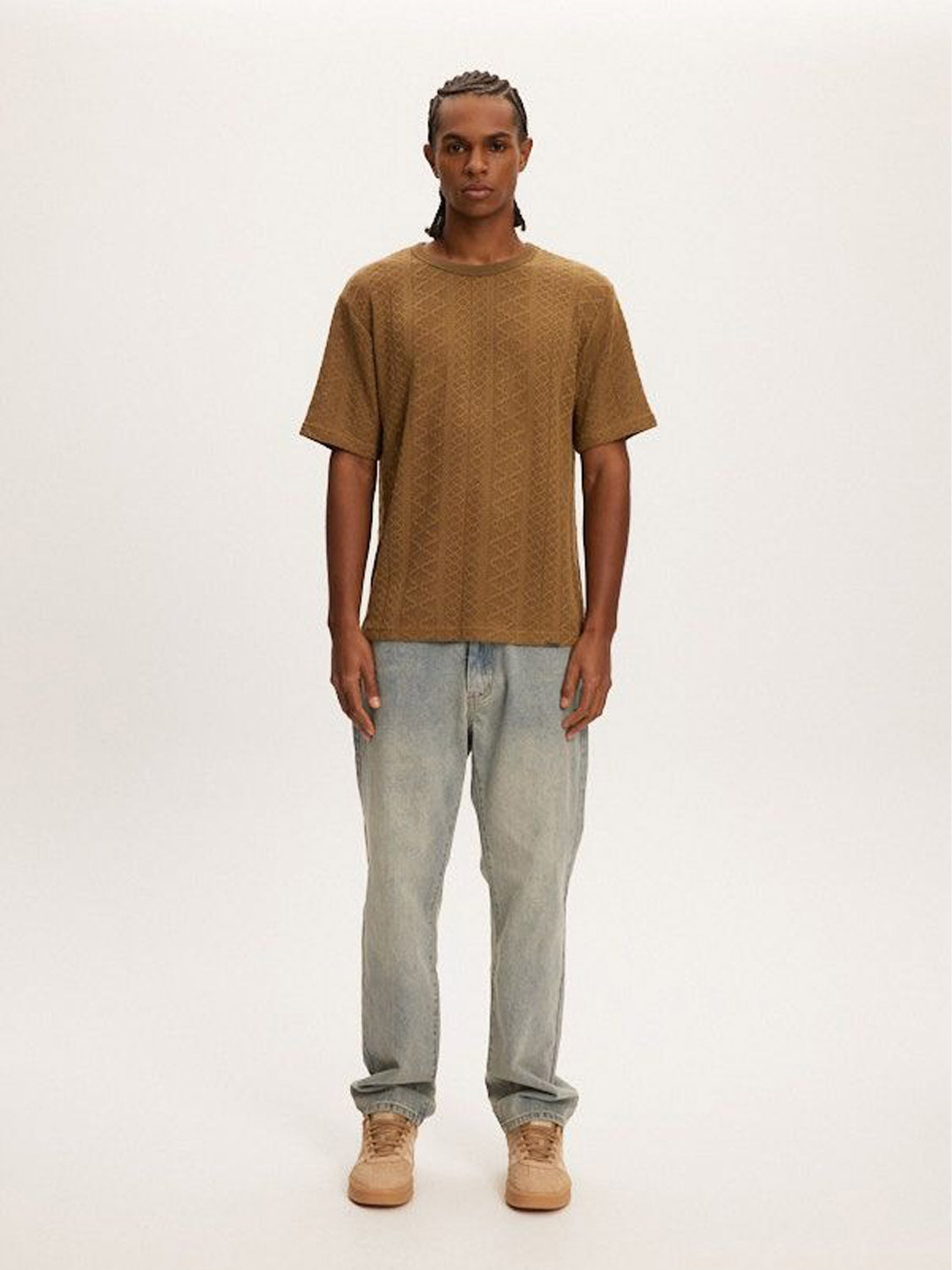 A person stands against a plain background wearing a Kuwalla Crochet Jersey Tee, light blue classic-fit jeans, and brown shoes. They have braided hair and a calm expression.