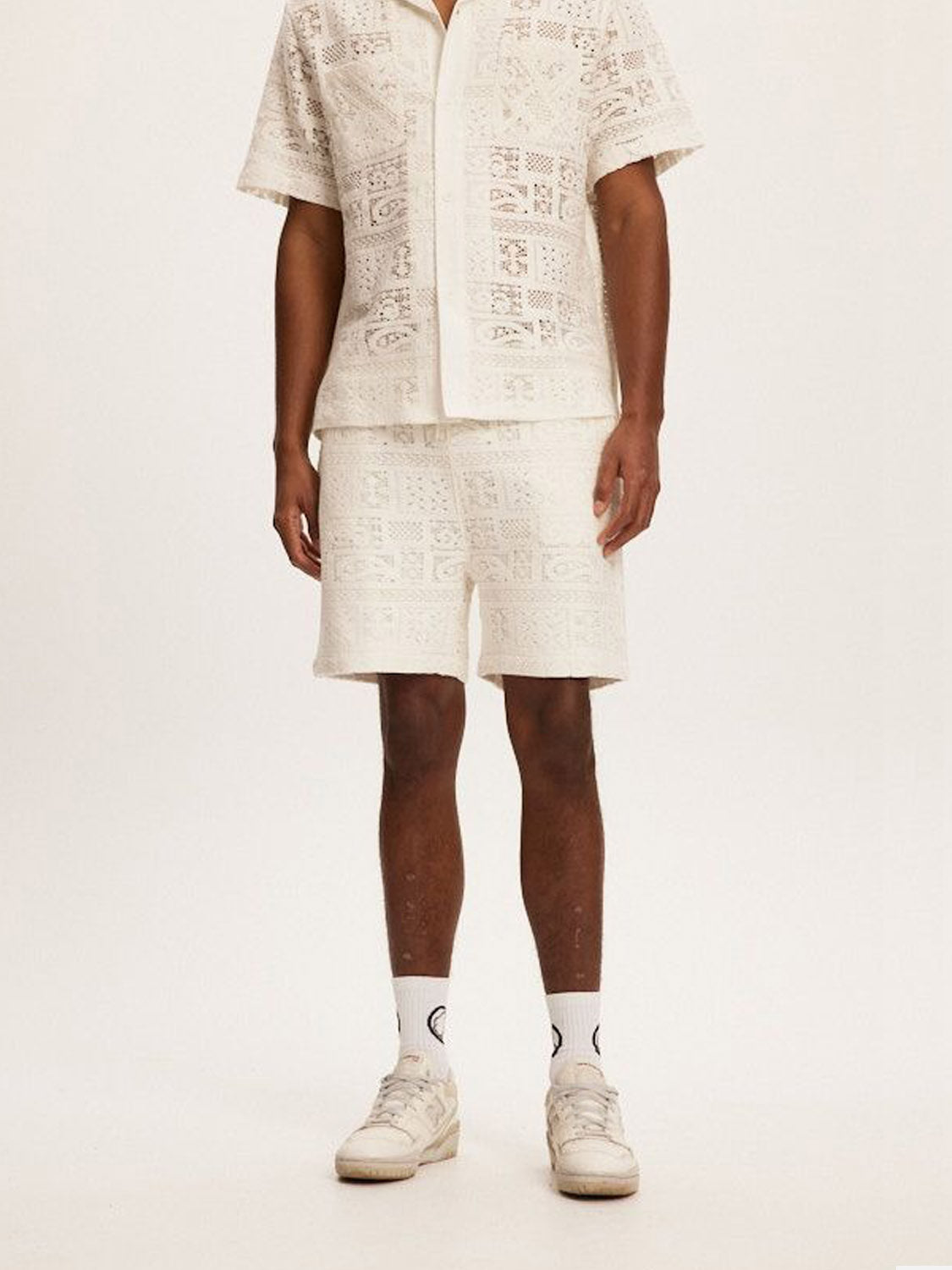 A person wears Kuwallas Crochet Short, a matching white lace-patterned short-sleeve shirt and shorts combo, designed with a relaxed fit ideal for summer days, paired with white socks and sneakers. The plain backdrop highlights the outfits texture.