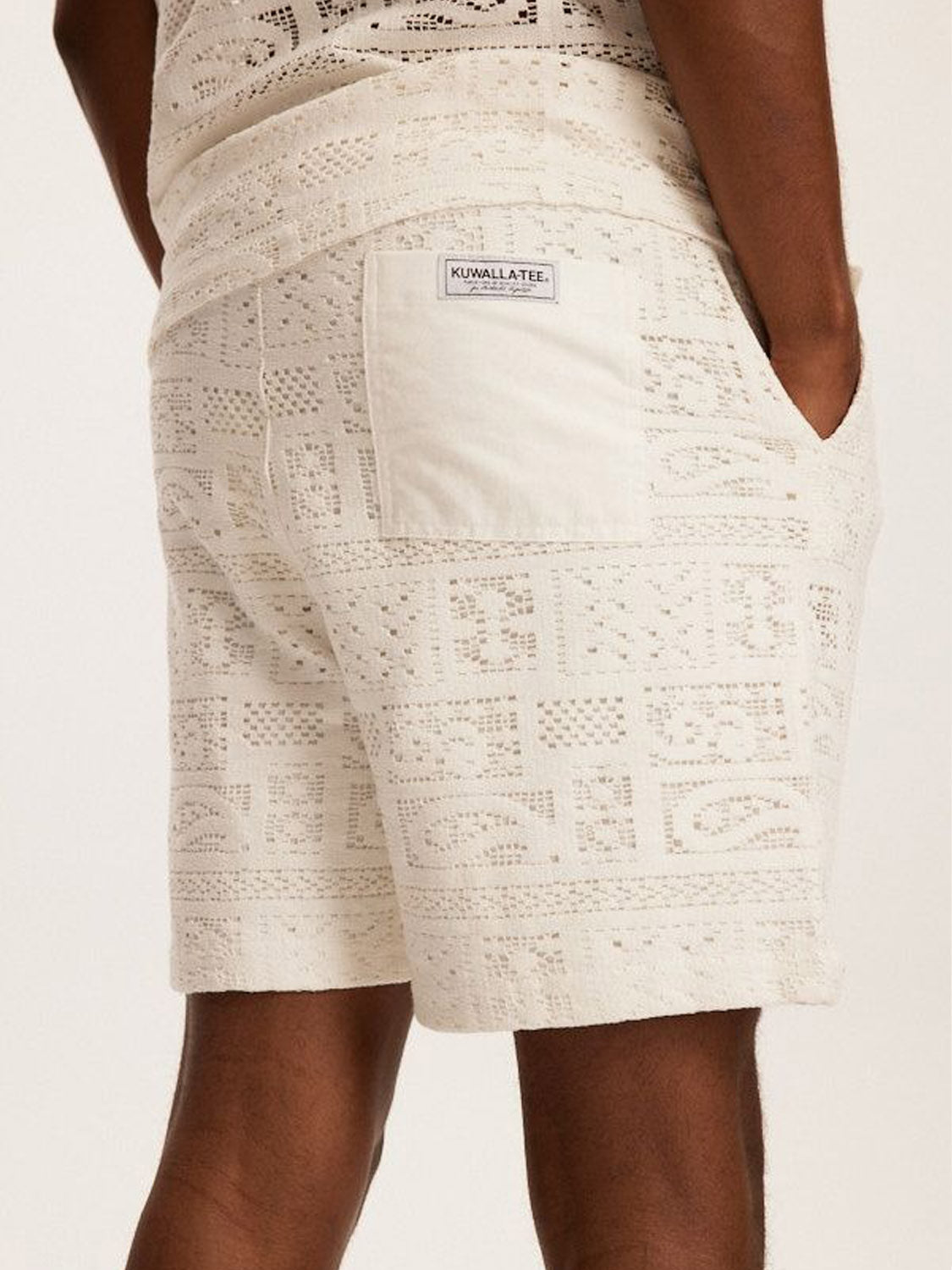 Someone wearing cream-colored Kuwalla Crochet Shorts with a relaxed fit, hands in pockets. Featuring geometric designs for a laid-back summer vibe, completed with a small label on the back pocket.