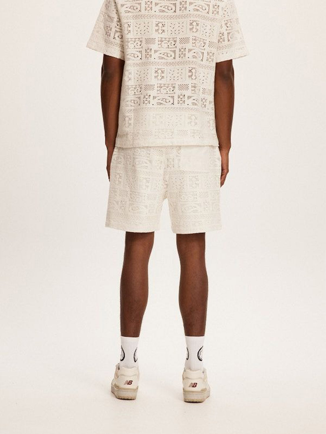 A person stands with their back turned, wearing a matching beige Kuwalla Crochet Short set in a relaxed fit. They pair it with white socks and sneakers featuring red accents. The crochet fabric suggests easy summer days, all set against a plain white backdrop.