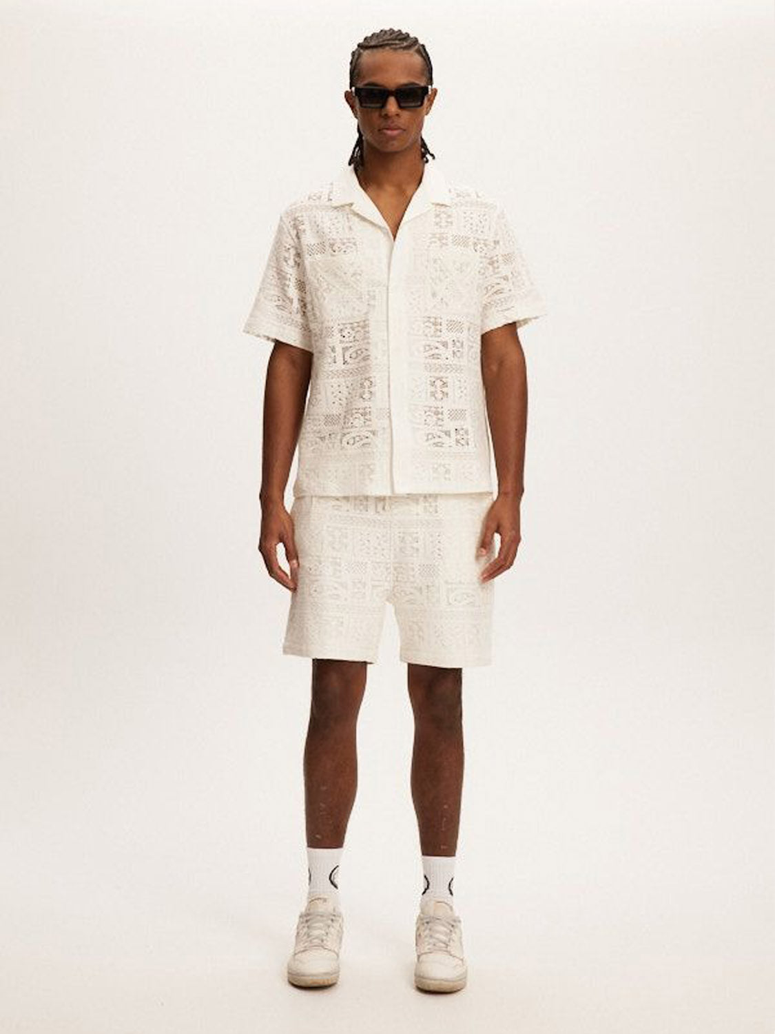 A person wearing sunglasses stands against a plain background, sporting Kuwallas stylish white crochet lace-patterned short-sleeve shirt and matching Kuwalla Crochet Short. The relaxed-fit ensemble is completed with white socks and sneakers, making it perfect for summer days.