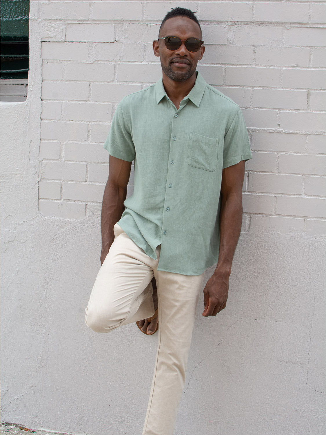 A man in ID Menswears Cruise Lightweight Stretch Pant wears sunglasses and a light green short-sleeve button-up shirt, standing casually with one leg bent against a light brick wall.