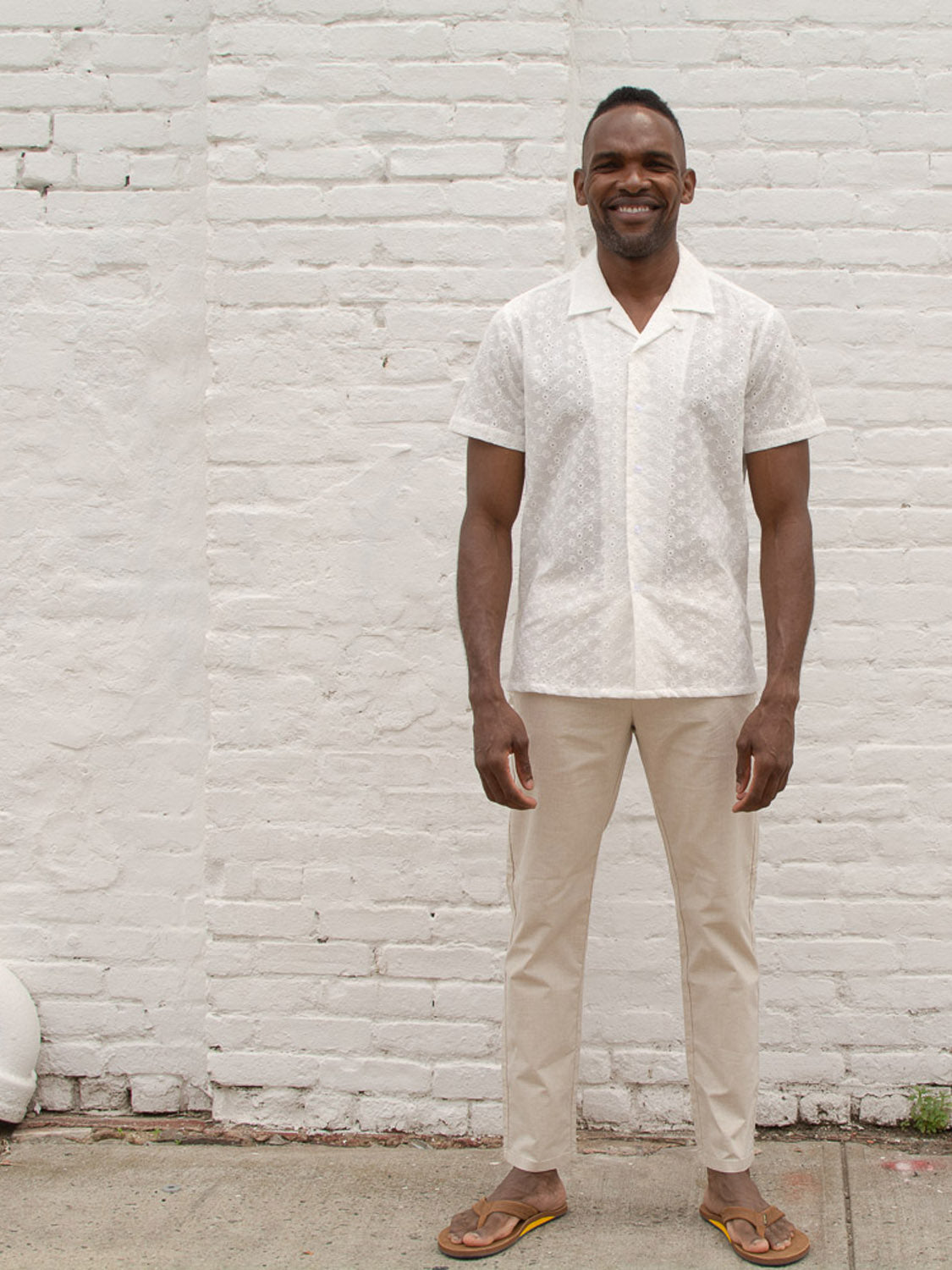 A man smiles in front of a white brick wall, wearing a white short-sleeve button-up shirt, brown flip-flops, and the Cruise Lightweight Stretch Pant by ID Menswear.