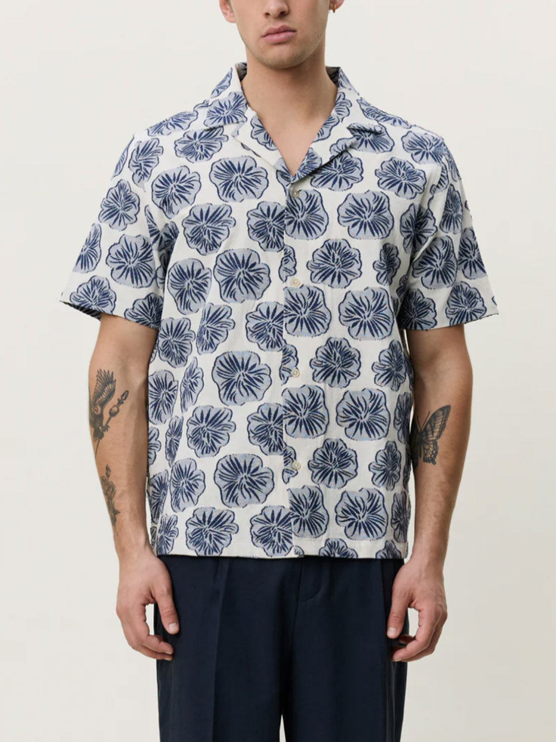 A man stands facing forward against a plain background wearing the Les Deux Dahlia Jacquard SS Shirt, a short-sleeve camp collar shirt with a blue and white pattern, paired with dark trousers; his arm tattoos are visible.