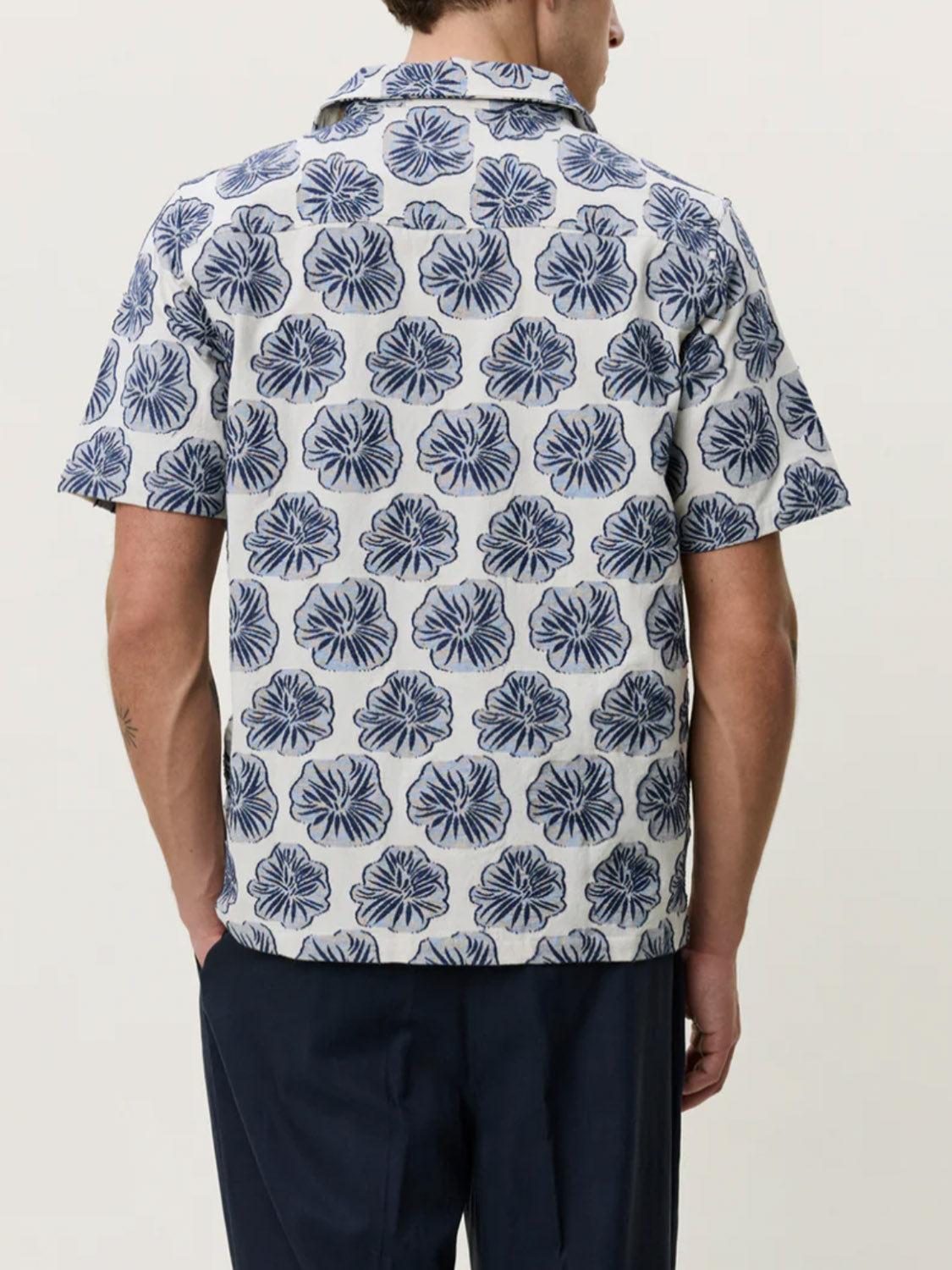 A man, seen from the back, wears the Les Deux Dahlia Jacquard SS Shirt with a blue and white floral design and short sleeves. He pairs it with dark trousers against a plain light background.