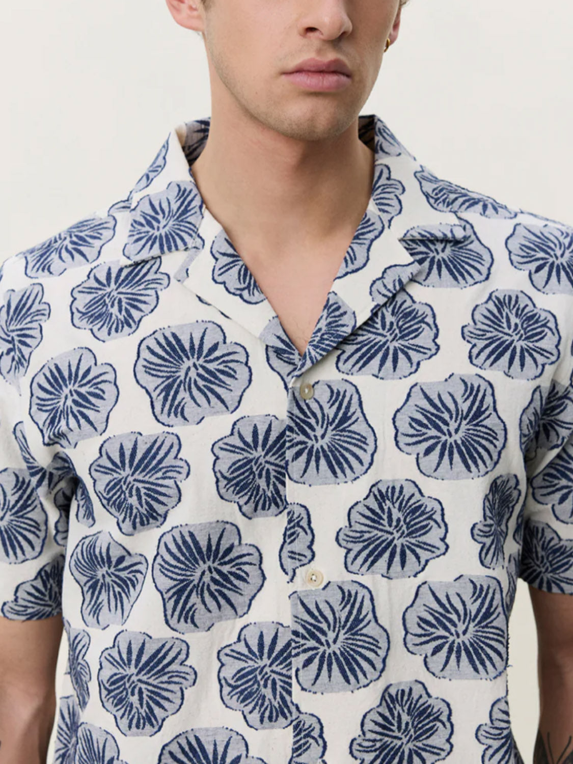 A person wearing the Les Deux Dahlia Jacquard SS Shirt, featuring a white camp collar and blue floral leaf print, is shown from shoulders to mid-torso against a plain light background.