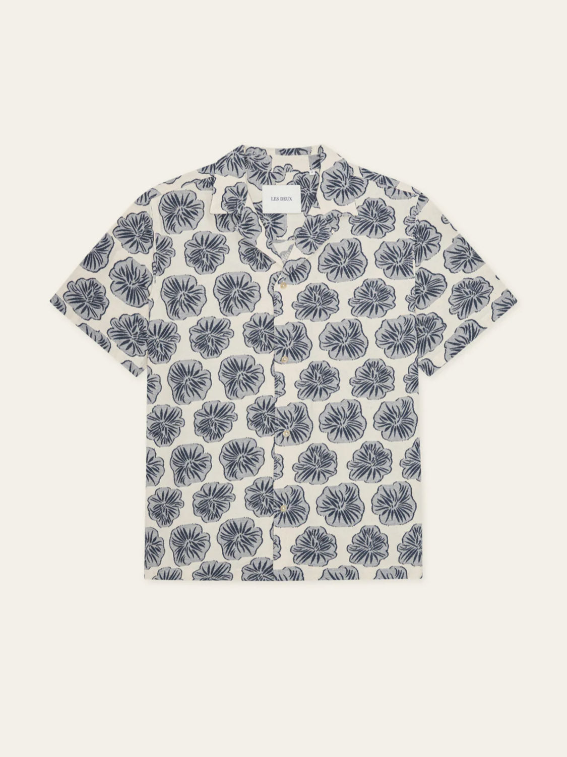 The Les Deux Dahlia Jacquard SS Shirt is a short-sleeved camp collar shirt with a white base, navy floral jacquard pattern, notched collar, and visible brand label inside the neckline.