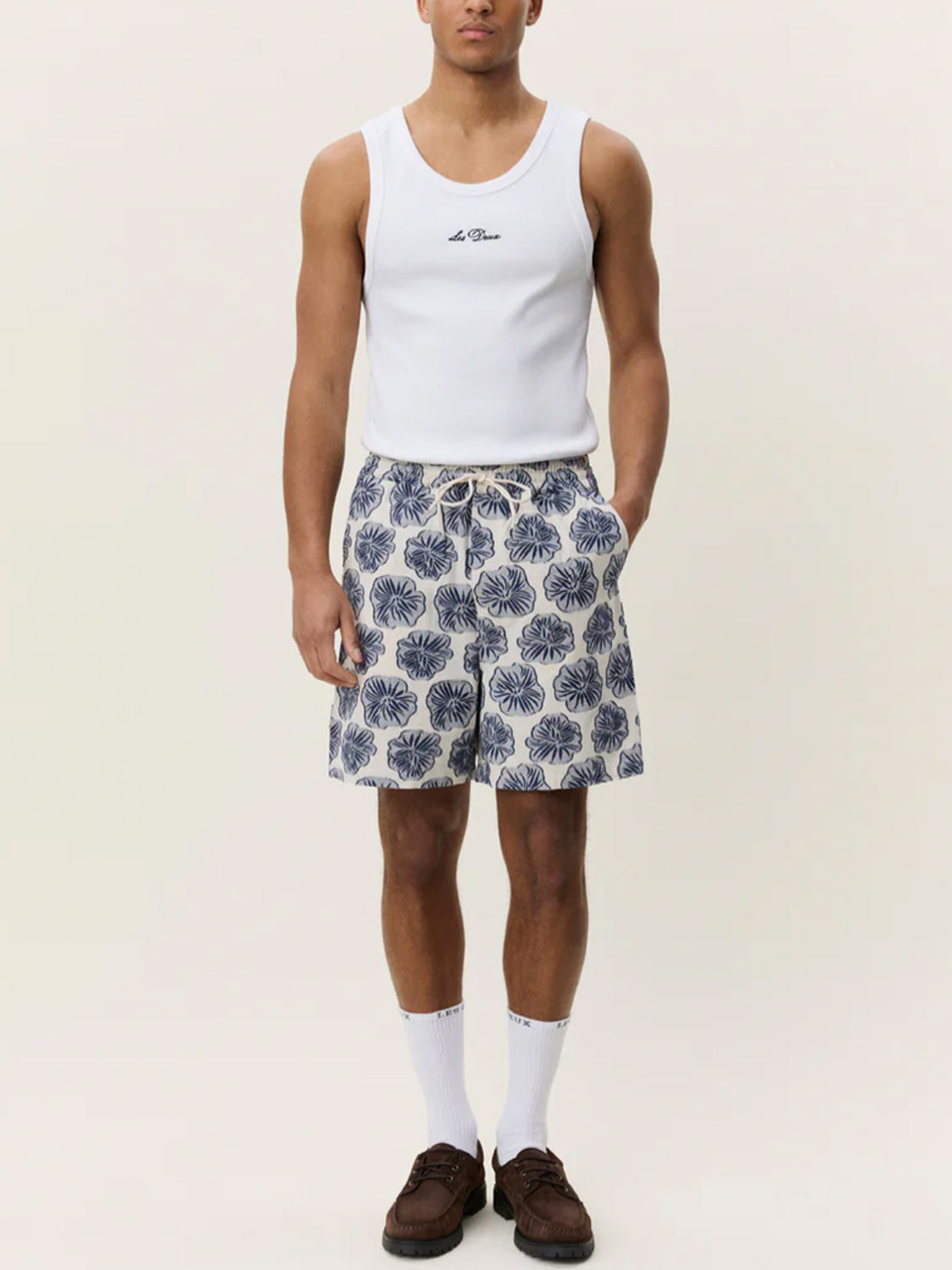 A man stands against a plain off-white background with his hands in his pockets, wearing a white tank top, Les Deux Dahlia Jacquard Shorts, white socks, and brown shoes.
