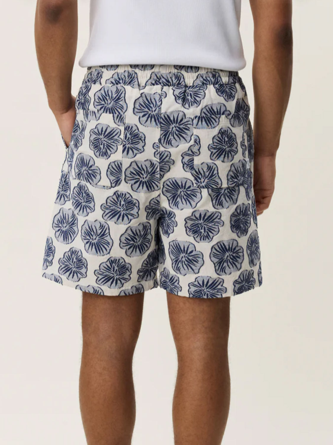 Wearing Les Deux's Dahlia Jacquard Shorts, a person in blue and white floral shorts with a drawstring waist and a white top stands facing away, hands in pockets.