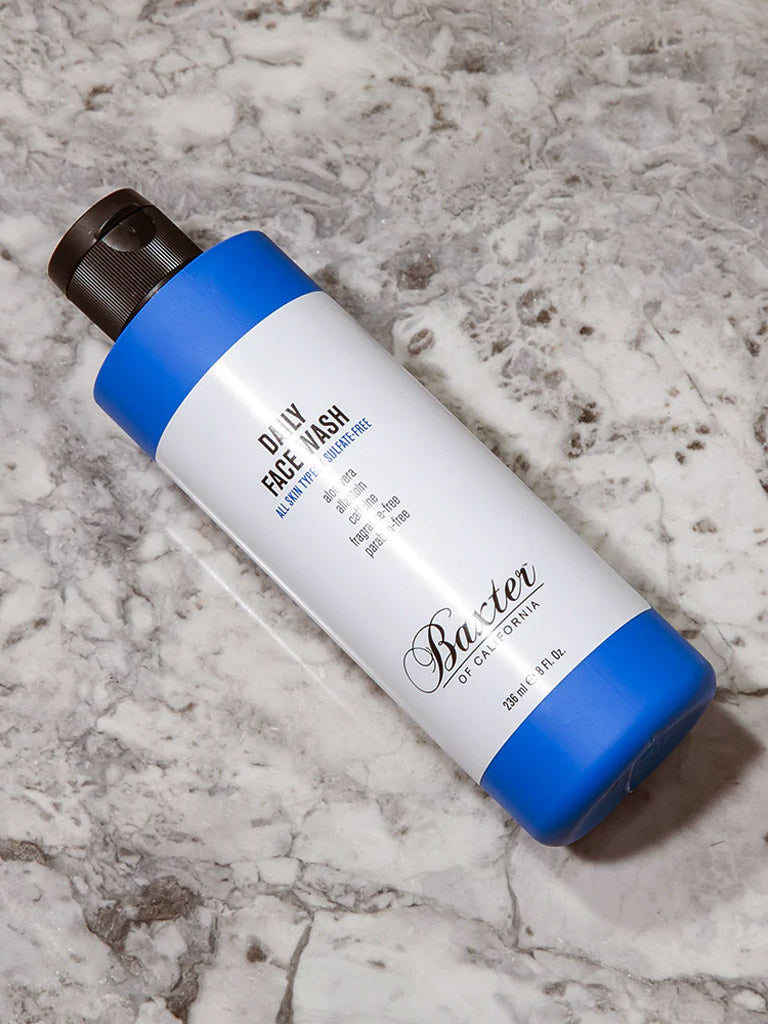 A bottle of Baxter Daily Face Wash, a gentle cleanser for sensitive skin, featuring blue ends and a black pump top, rests on a gray and white marble surface.