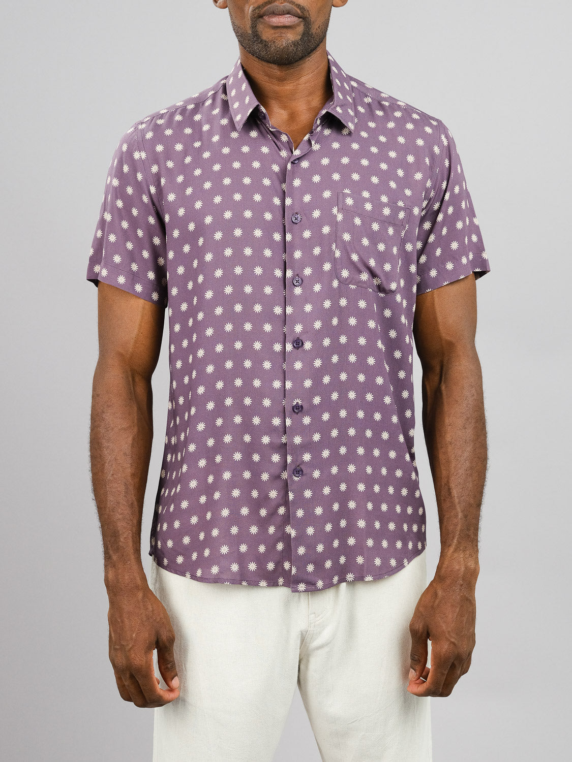 A man wears the Daze Printed Rayon Shirt by ID Menswear—a short-sleeve, purple shirt with white circular dots—paired with white pants against a grey background; perfect for warm weather and fans of bold prints.