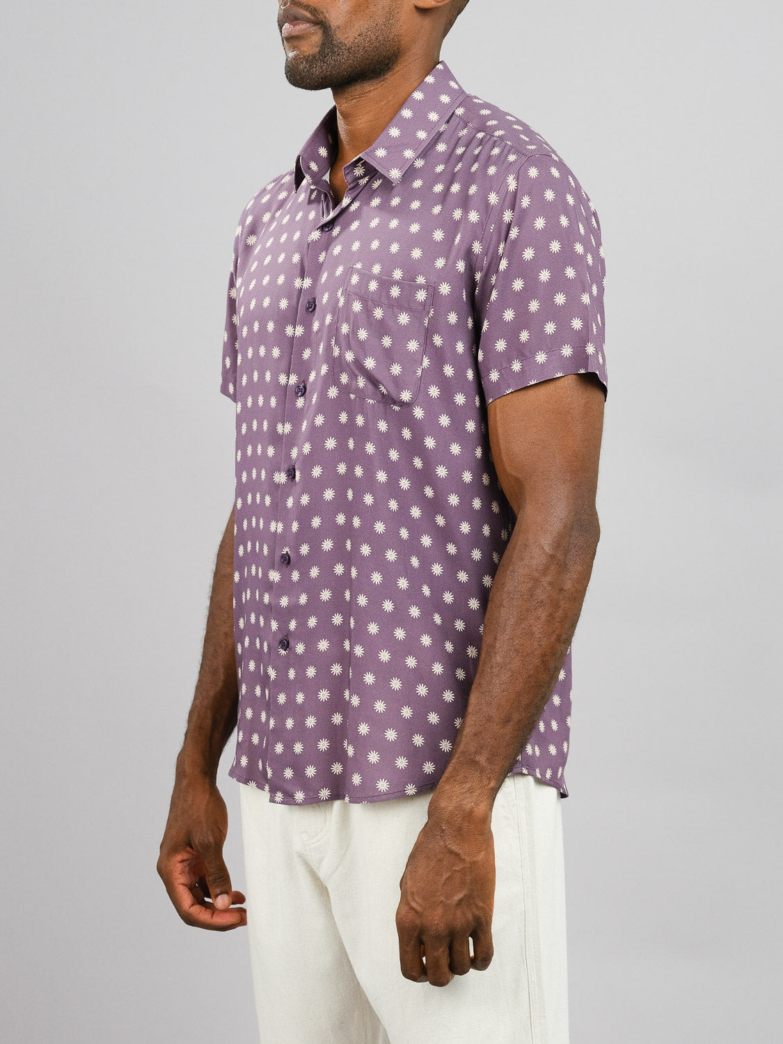 A man wears the Daze Printed Rayon Shirt by ID Menswear, featuring a purple and white dot pattern, paired with light pants against a gray background—ideal for warm weather.