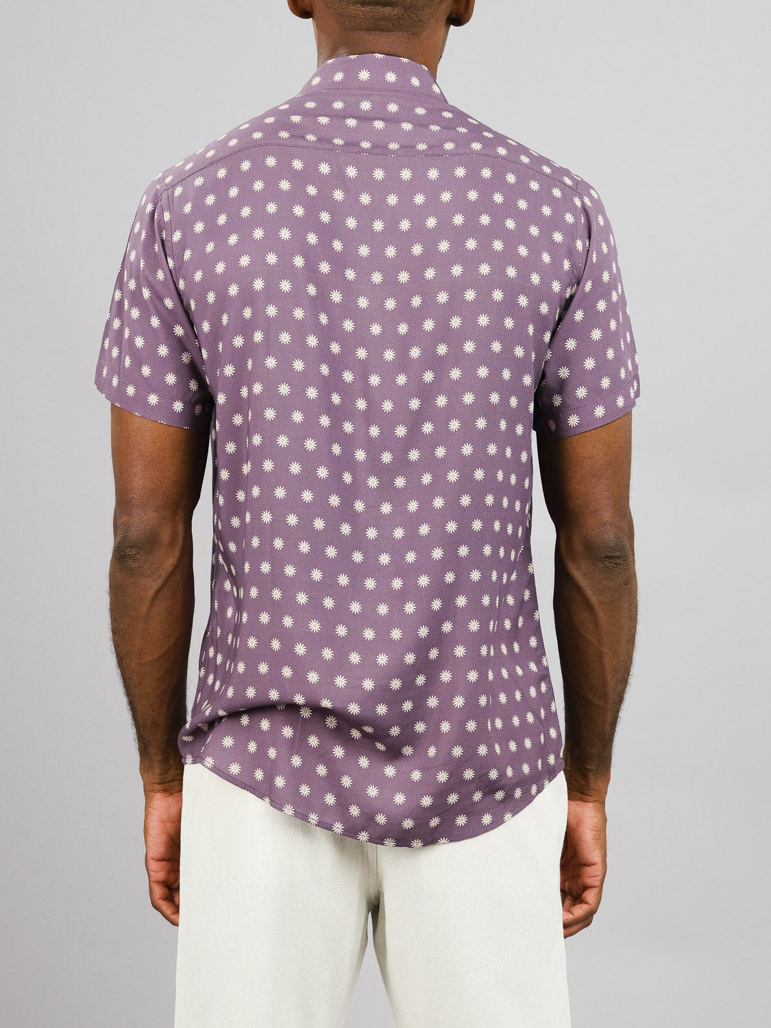 A person seen from behind wears the ID Menswear Daze Printed Rayon Shirt, a short-sleeve purple top with white polka dots, paired with off-white pants—ideal for warm weather—standing against a plain gray background.