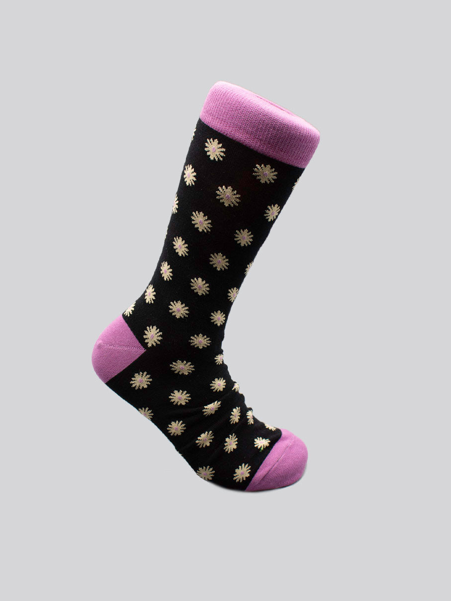 The Dazed Graphic Socks by ID Menswear feature black fabric with white daisy patterns and pink accents on the toe, heel, and cuff. Made from high-quality combed cotton, theyre a modern essential—shown here against a light gray background.
