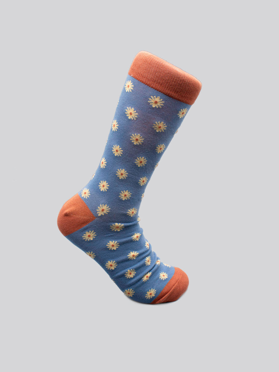 The Dazed Graphic Socks by ID Menswear feature blue combed cotton with orange heel, toe, and cuff, detailed with small white and yellow flower motifs—a bold statement piece for modern wardrobes, shown on a plain light gray background.