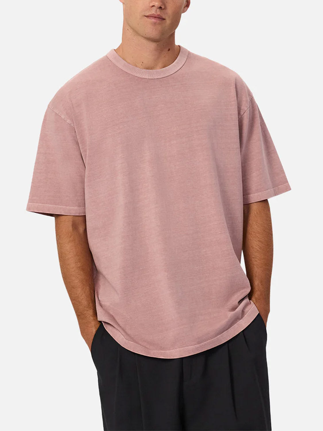 A person stands with hands in pockets against a plain light background, wearing Industrie’s The Del Sur Tee—a relaxed fit, short-sleeve, light pink crew neck t-shirt—and dark pants.