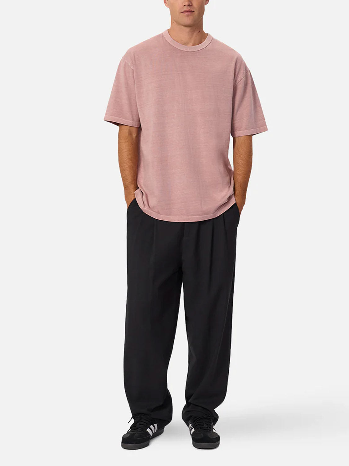 A person stands with hands in pockets, wearing The Del Sur Tee by Industrie—a relaxed fit, light pink crew neck—paired with wide black pants and black Adidas sneakers, against a plain white background.