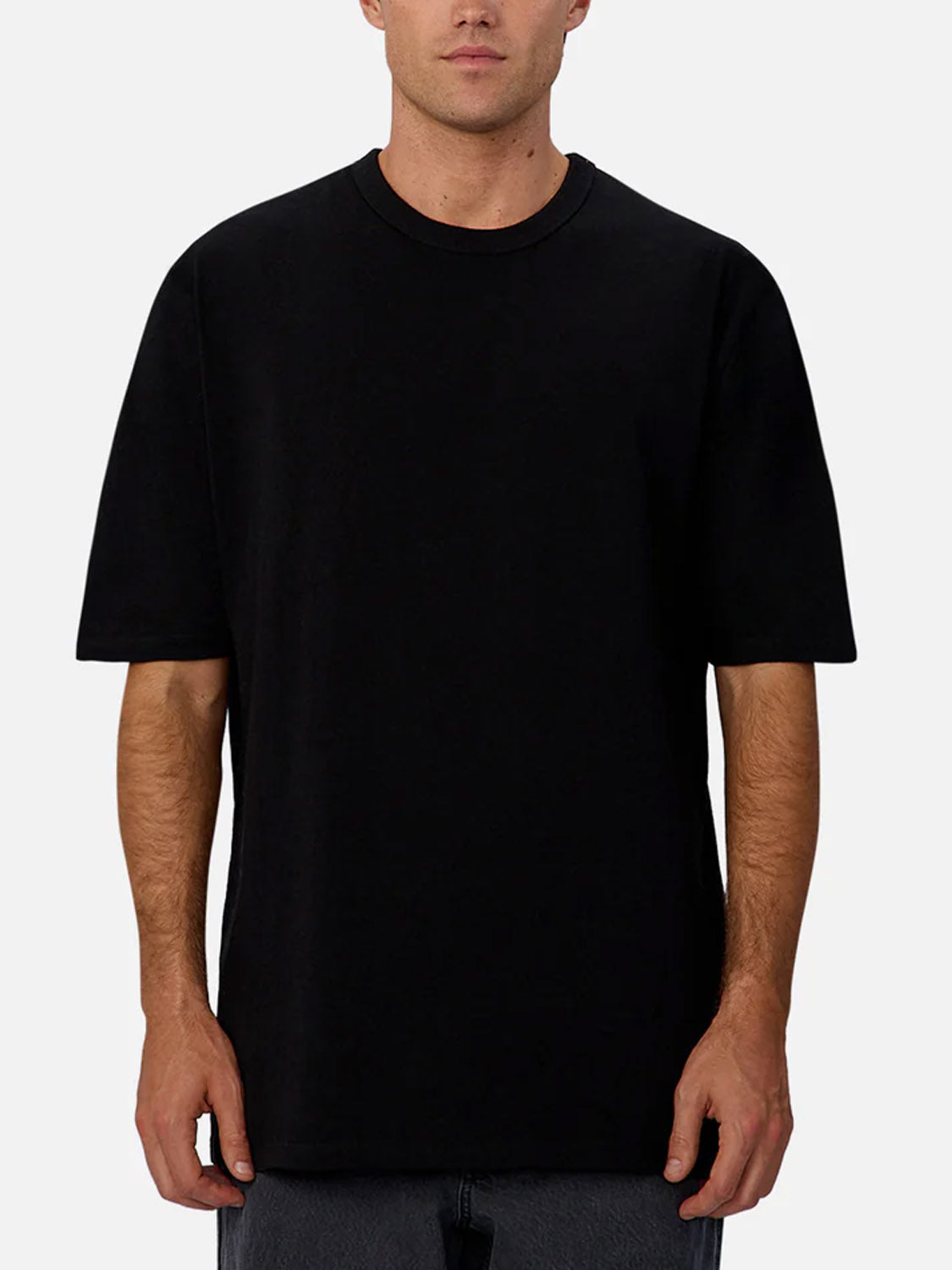 A man stands facing forward against a light gray background, wearing Industrie’s The Del Sur Tee—a plain black, relaxed fit crew neck t-shirt made from 100% cotton—paired with dark pants. His face is partially visible.