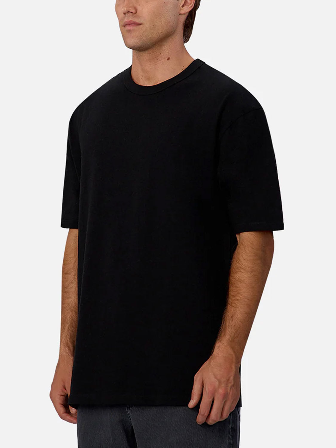 A man in Industrie’s The Del Sur Tee, a plain black 100% cotton crew neck, and dark pants stands against a light gray background, facing slightly to the side.