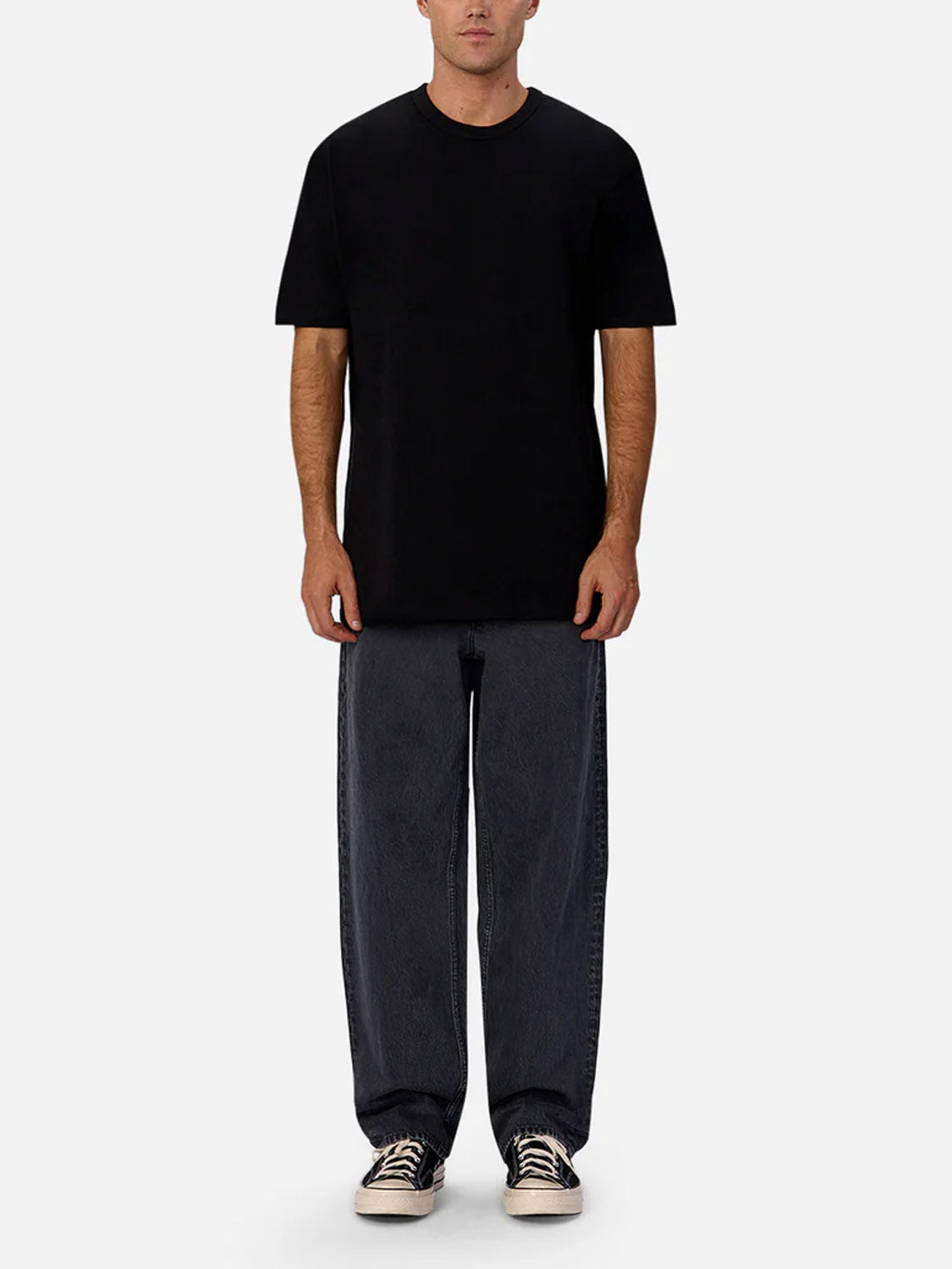 A man faces forward wearing The Del Sur Tee by Industrie, a relaxed fit 100% cotton black crew neck, paired with loose dark gray jeans and black-and-white sneakers against a light gray background.