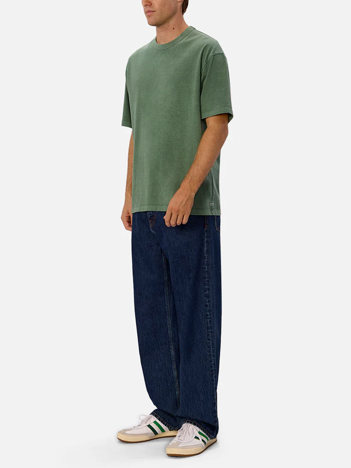 A person stands slightly sideways in The Del Sur Tee by Industrie, a relaxed-fit 100% cotton crew neck in green, paired with wide-leg blue jeans and white sneakers with green and black stripes, against a plain light background.