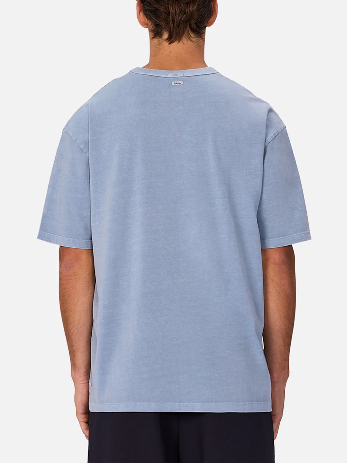 A person with short brown hair, seen from behind, wears the Industrie Del Sur Tee in light blue paired with black shorts against a plain light background.