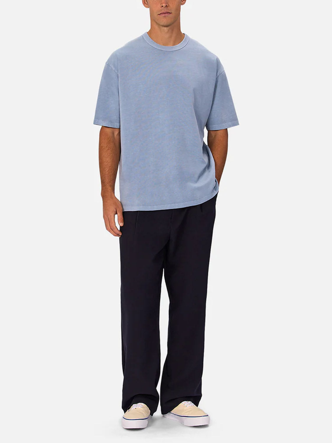 A man stands against a plain background wearing Industrie’s The Del Sur Tee in light blue, relaxed fit, paired with loose dark pants and beige sneakers. His hands are by his sides and in his pocket.
