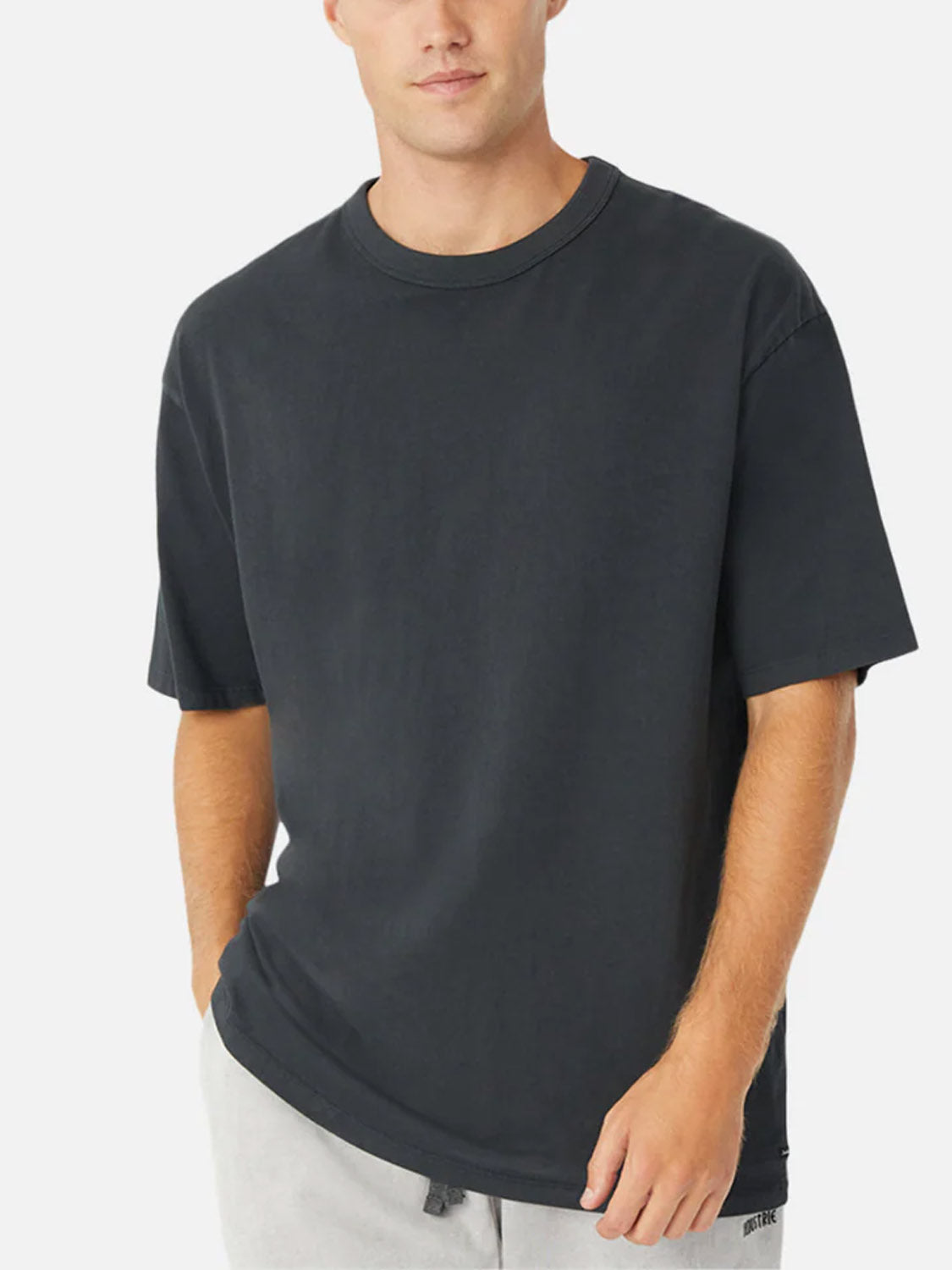 A man wears the Industrie Del Mar Boxy T-Shirt, a relaxed fit heavyweight cotton tee in dark gray, paired with light gray pants, standing with one hand in his pocket against a plain light background.
