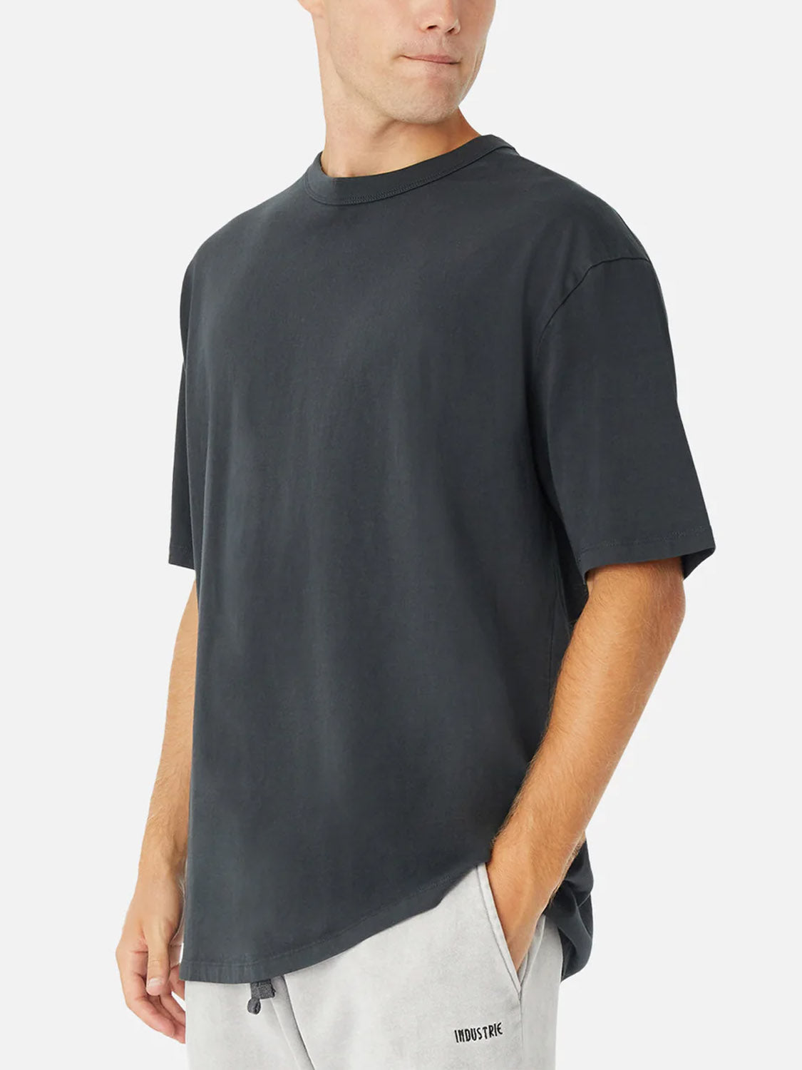 A man wears the Industrie Del Mar Boxy T-Shirt in black and light gray shorts with “INDOSYNC” printed on the left, his left hand in his pocket, standing against a plain white background.