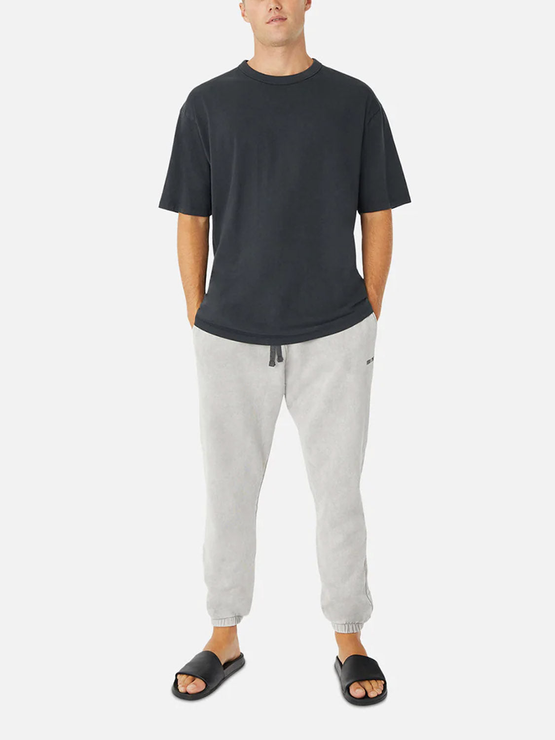 A person stands with hands in pockets, wearing the Industrie Del Mar Boxy T-Shirt, relaxed fit heavyweight cotton, paired with light gray jogger pants and black slide sandals against a plain white background.