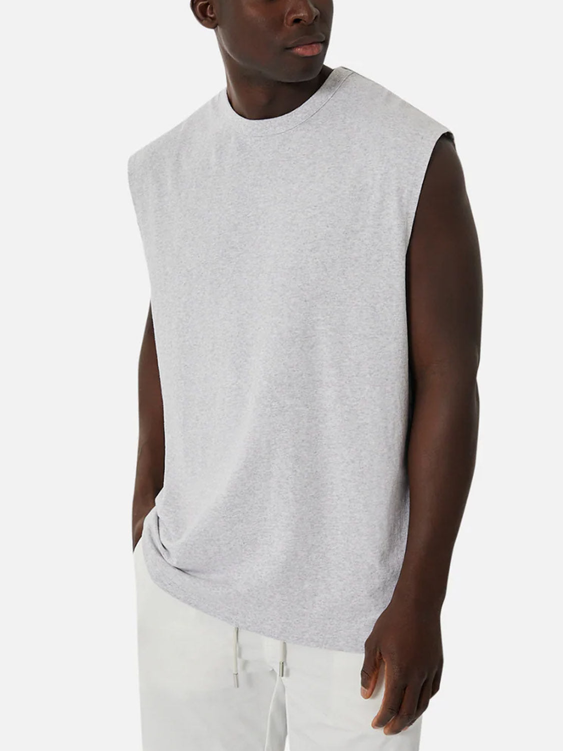 A man models the Industrie Del Sur Sleeveless T-Shirt in light gray paired with white pants, standing with his left hand in his pocket and looking to his left against a plain white background.