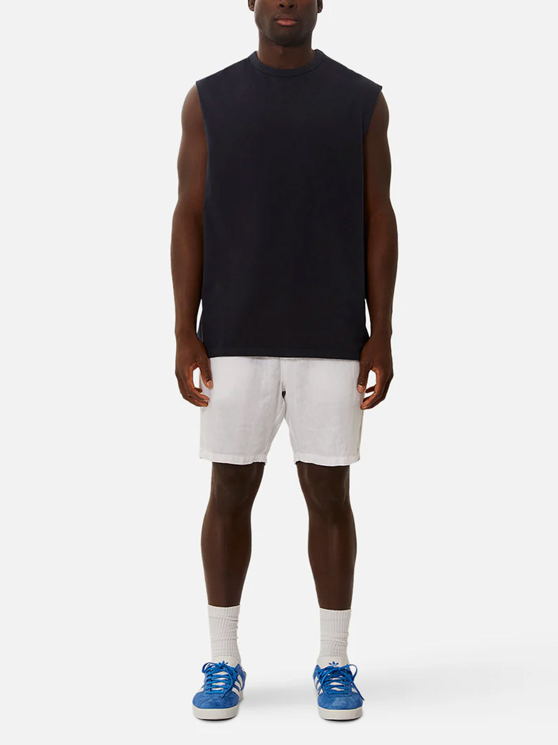 A person stands facing forward against a plain white background, wearing an Industrie Del Sur Sleeveless T-Shirt, white shorts, white socks, and blue sneakers.