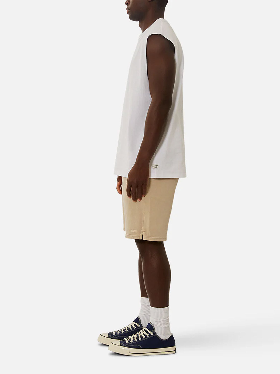 A man stands in profile wearing the Industrie Del Sur Sleeveless T-Shirt, beige shorts, white crew socks, and black sneakers with white soles against a plain light background.